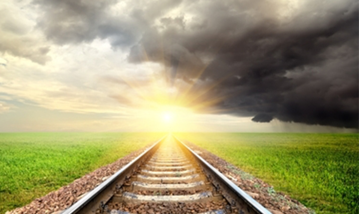 Railway tracks leading into a bright sunrise with dark storm clouds on the right and green fields on both sides.