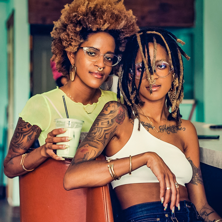 Two women with glasses, tattoos, and jewelry. One woman is holding a clear cup with a green beverage.