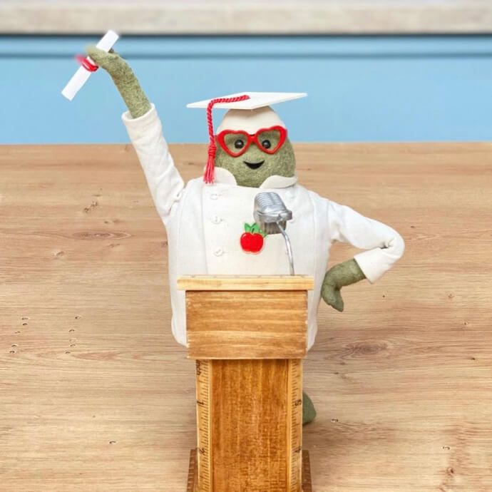 Stuffed green figure wearing red glasses, white chef coat, and graduation cap, holding a diploma behind a podium.