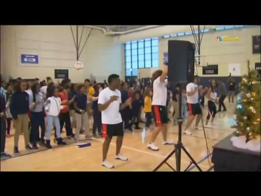 People exercise in a gymnasium; a group in the background follows the instructors in the foreground.