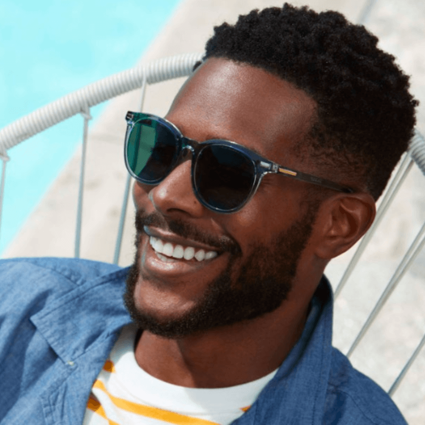 Man wearing round, blue-tinted sunglasses and a blue shirt, smiling brightly.