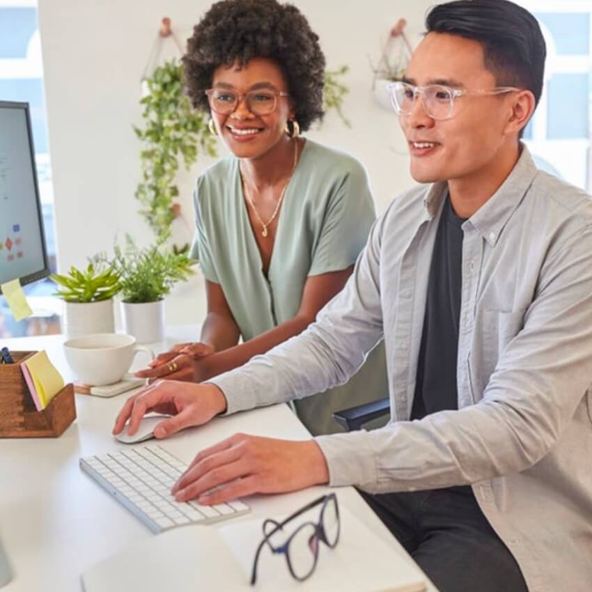 Two people wearing glasses sitting at a desk with a keyboard, plants, glasses case, and white coffee cup.