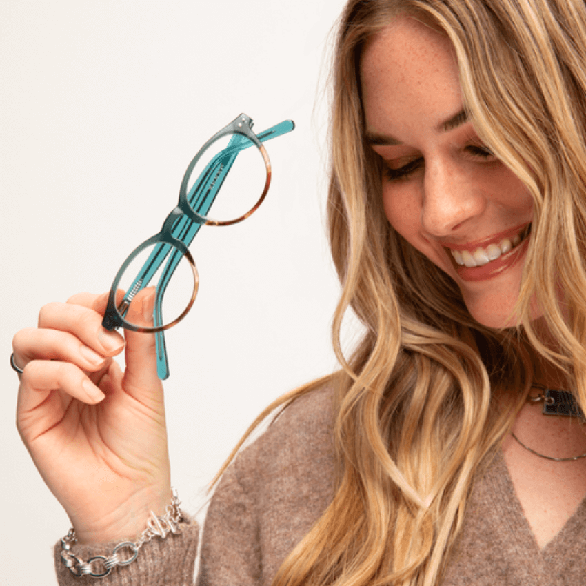 Woman holding turquoise round eyeglasses with gold rims, smiling.