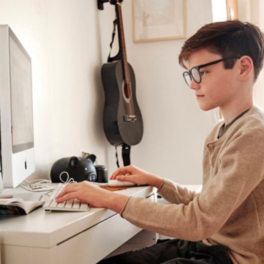 Boy typing on a white computer keyboard with a gray monitor on a white desk.