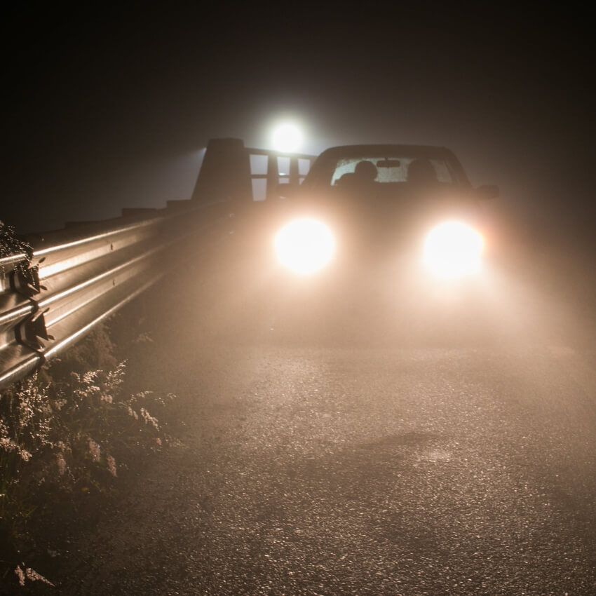 Car with headlights on in a foggy environment near a metal guardrail.