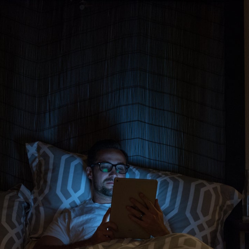 Person in bed wearing glasses, illuminated by a tablet screen. Hexagonal pattern on pillows and bedding.