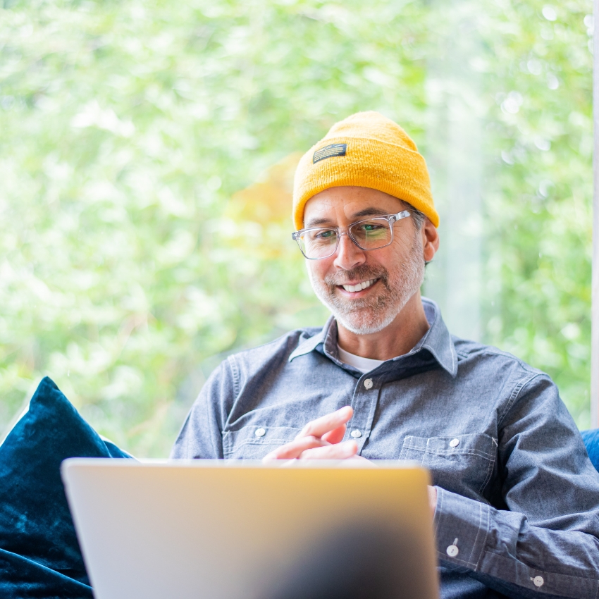Man wearing a yellow beanie, glasses, and a gray button-up shirt, smiling while using a laptop.