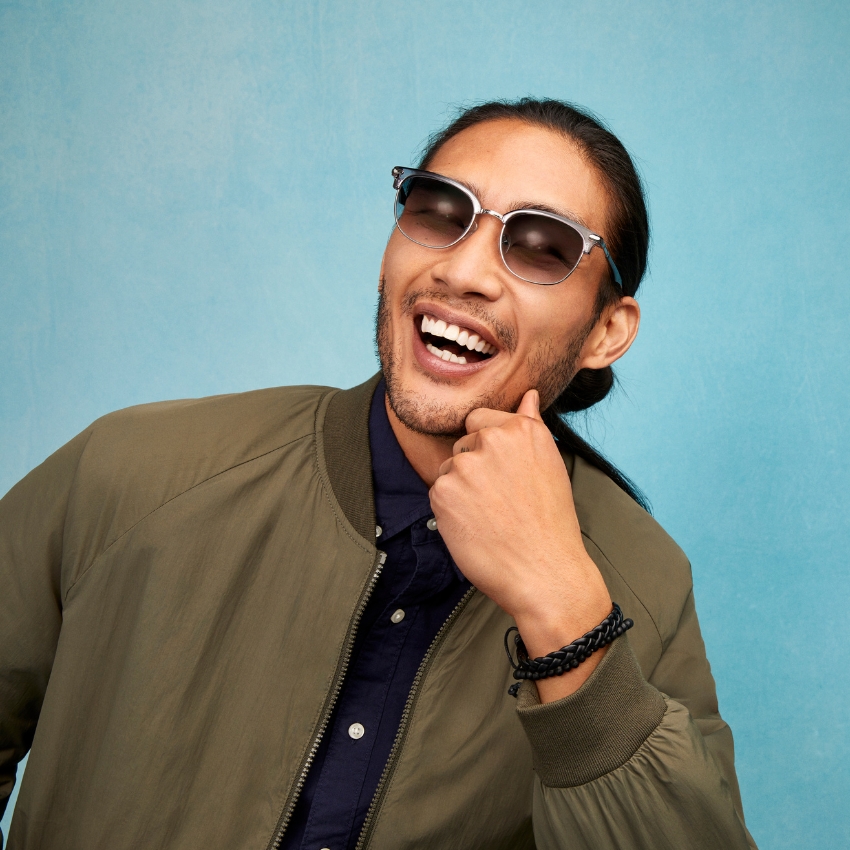 Man wearing reflective sunglasses, a dark shirt, and an olive green jacket, smiling broadly.