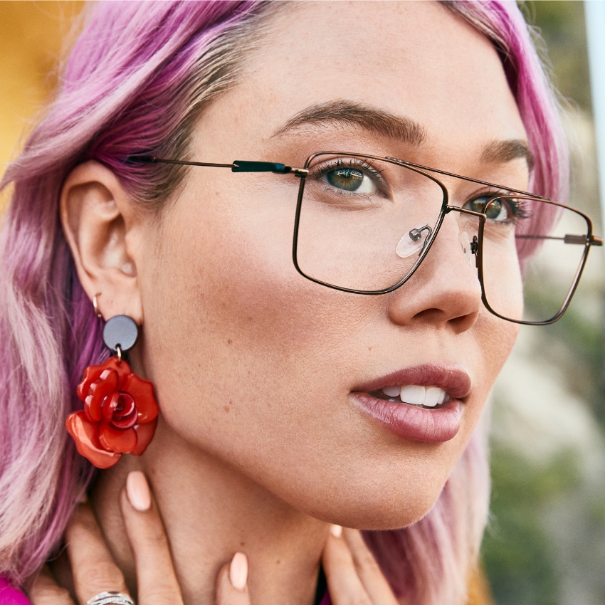 Rectangular glasses with thin metal frame and clear lenses worn by a person with pink hair and red rose earrings.