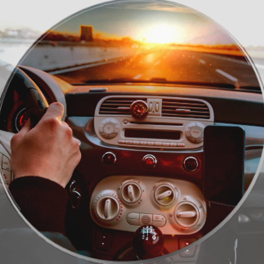 View of a car dashboard with a hand gripping the steering wheel, driving towards a sunset on an open road.