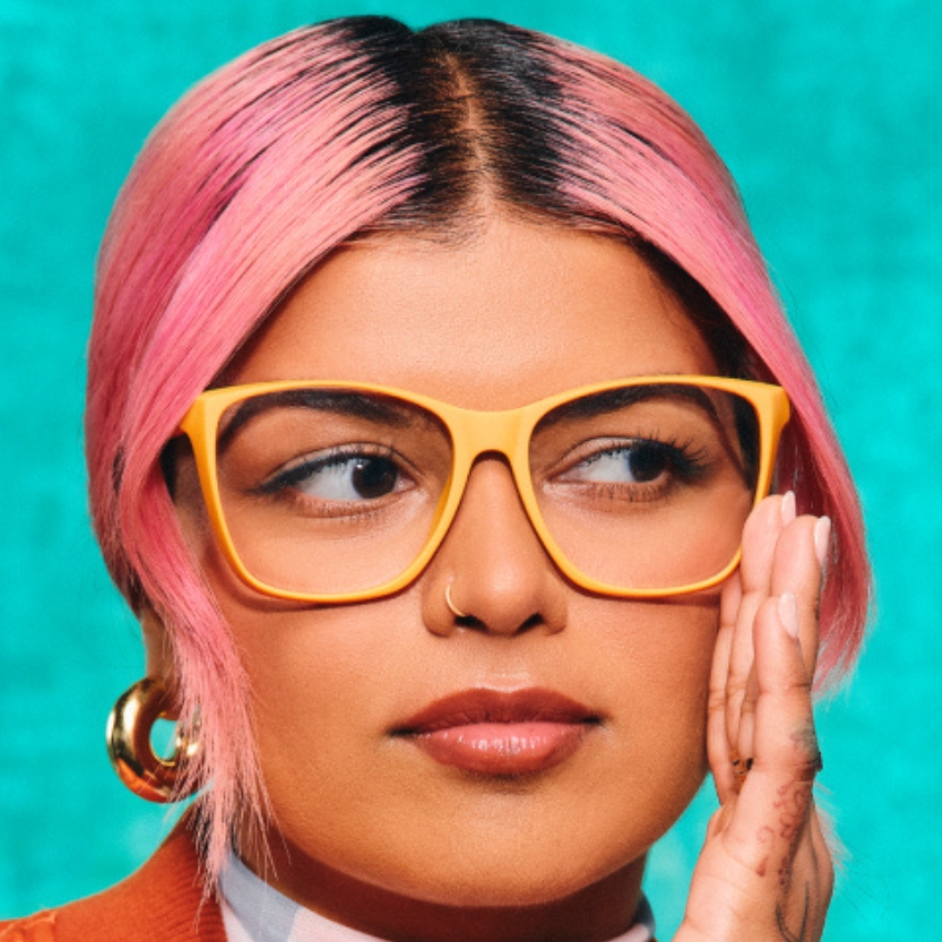 Person wearing oversized, bright yellow eyeglass frames, gold hoop earrings, and sporting pink hair.