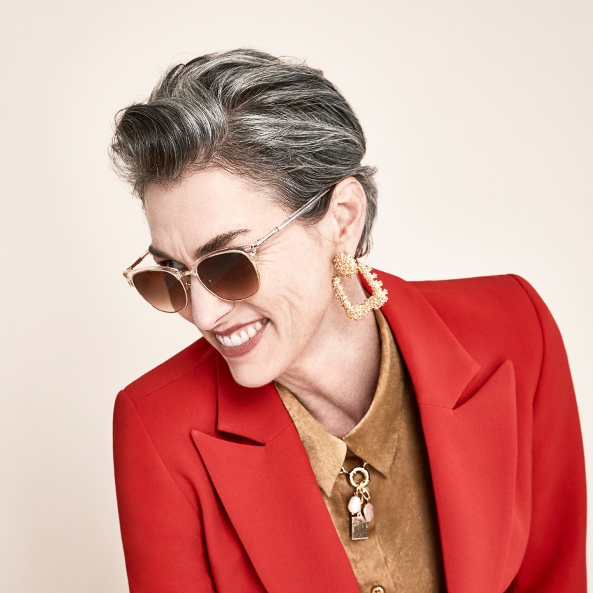 Woman wearing gold-framed sunglasses, large gold earrings, red blazer, and brown shirt with gold details.