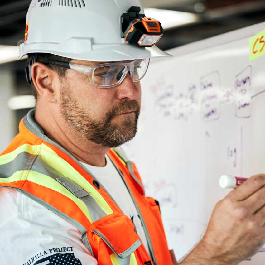 Construction worker wearing a hardhat with a headlamp, safety glasses, and a high-visibility vest writing on a whiteboard.