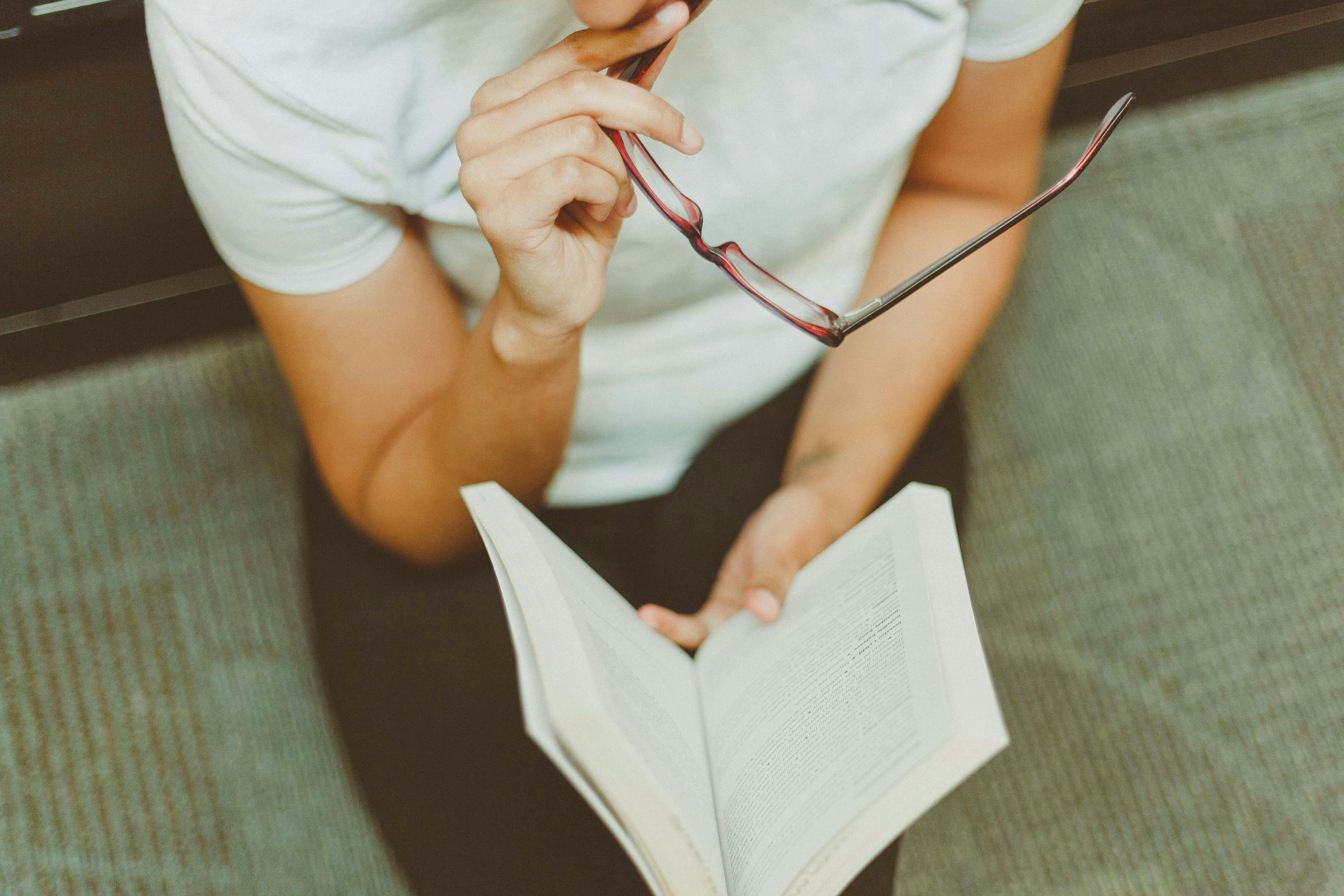 Person reading a book while holding glasses near mouth.