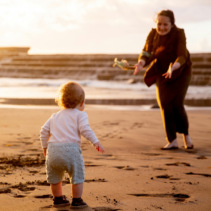 Toddler in blue shorts and white top walking on the beach towards an adult with open arms.