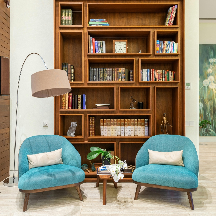 Teal chairs with white pillows, wooden coffee table with plant, and a large wooden bookshelf filled with books.