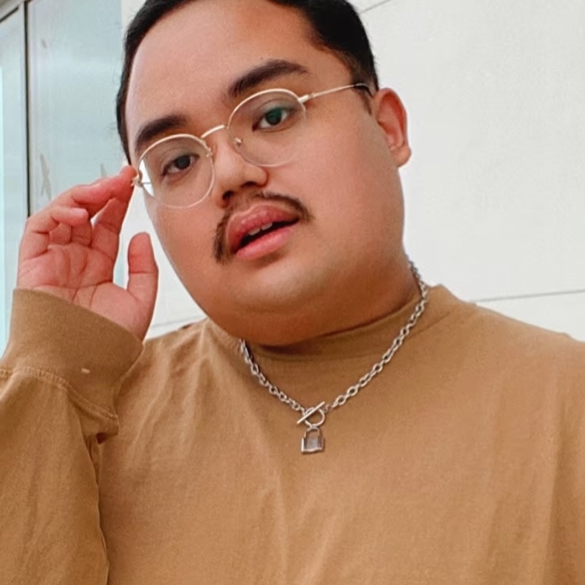 Person wearing clear glasses, a brown shirt, and a silver chain necklace with a lock pendant.