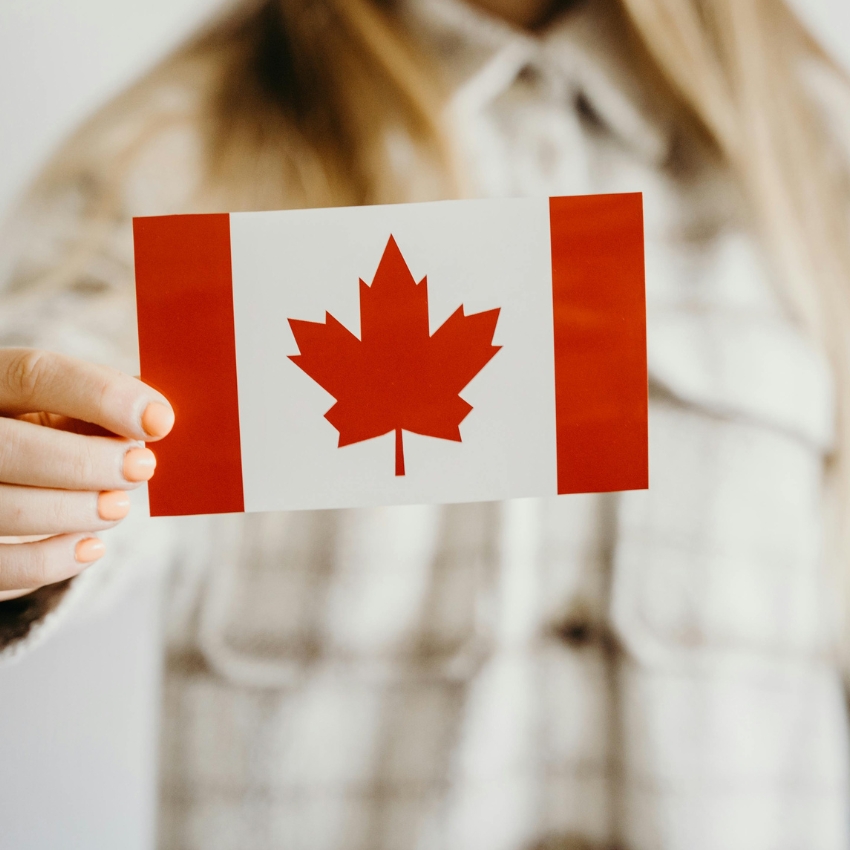 Hand holding a small Canadian flag featuring a red maple leaf in the center with red borders on the sides.