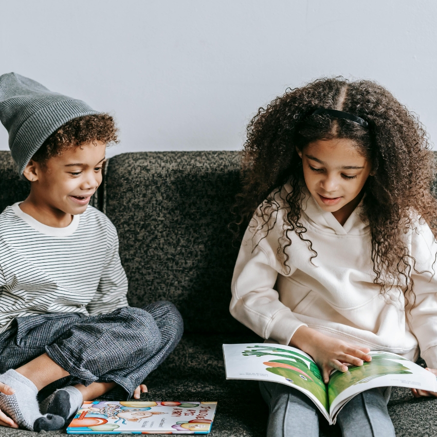 Two children reading illustrated books while sitting on a gray sofa.