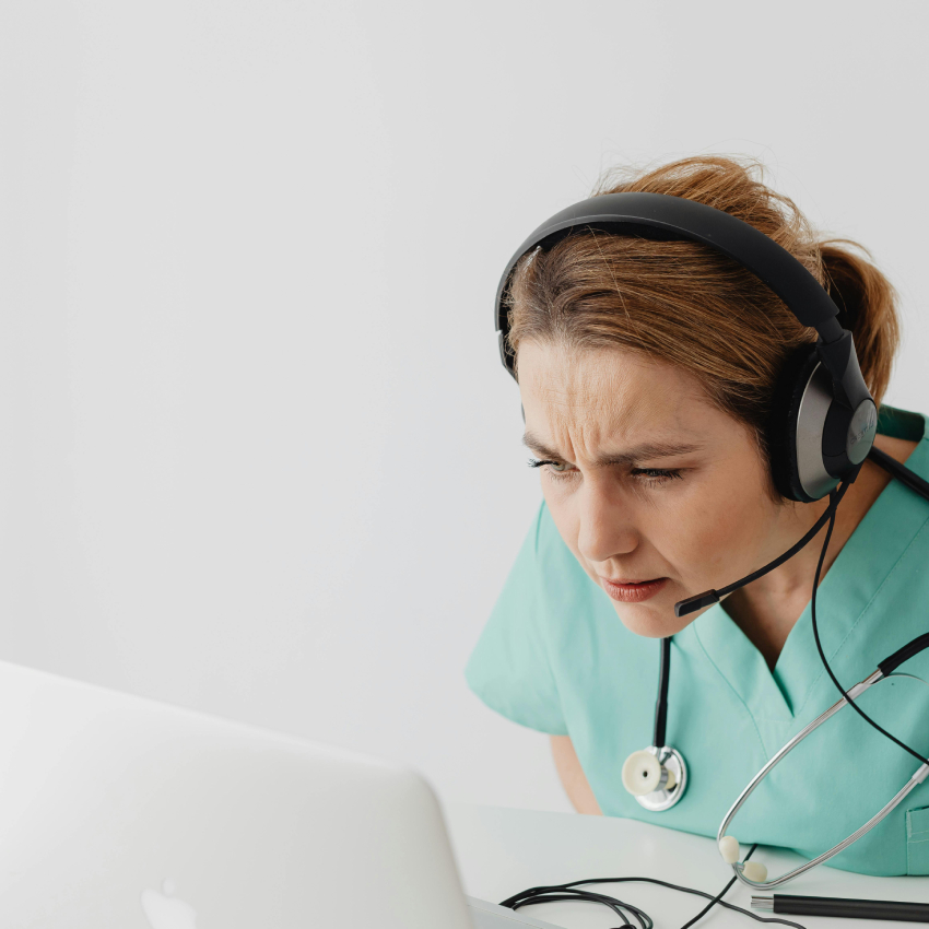 Nurse in scrubs wearing a headset and stethoscope, looking intently at a laptop screen.